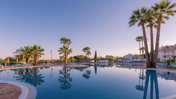 A clear swimming pool surrounded by palm trees and lounge chairs, with a building in the background. - Apartment 'Golden Sky' with Shared Pool, Wi-Fi and Air Conditioning (Cabanas de Tavira)