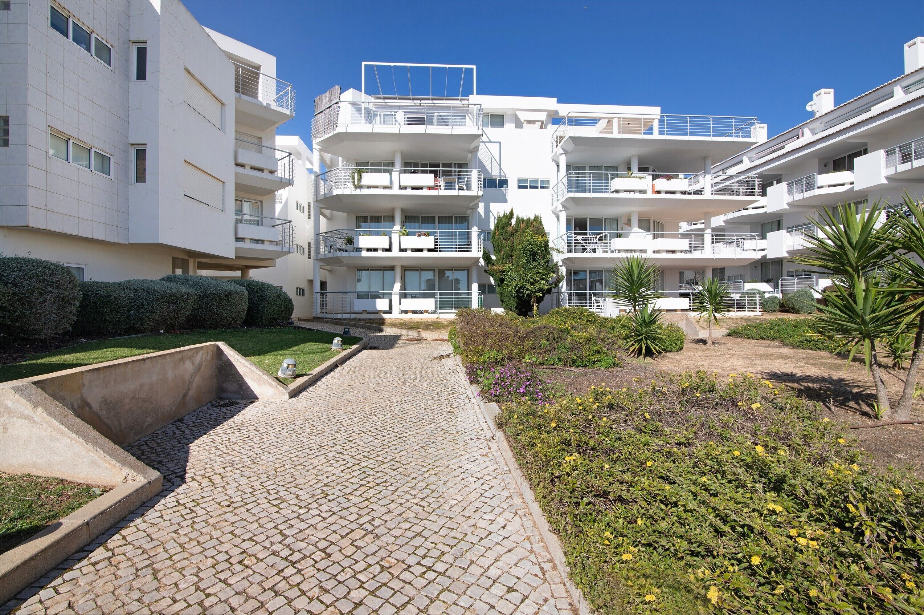 Un moderno edificio residencial de varios pisos con balcones, un patio empedrado y jardines ajardinados.