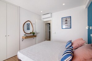 A bedroom with a bed, pillows, a wall-mounted mirror, a framed picture, and a small shelf with a plant.