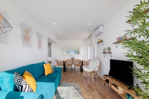 A living room with a teal sofa, a dining table with chairs, a TV on a wooden stand, and a large plant.