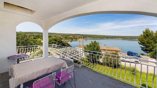 Un balcon avec une table et des chaises, donnant sur un lac et une route bordée de voitures garées.