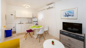 Un espace de vie compact comprenant un coin cuisine, un espace repas avec une table ronde et des chaises, un téléviseur posé sur un meuble en bois et une œuvre d’art encadrée accrochée au mur.
