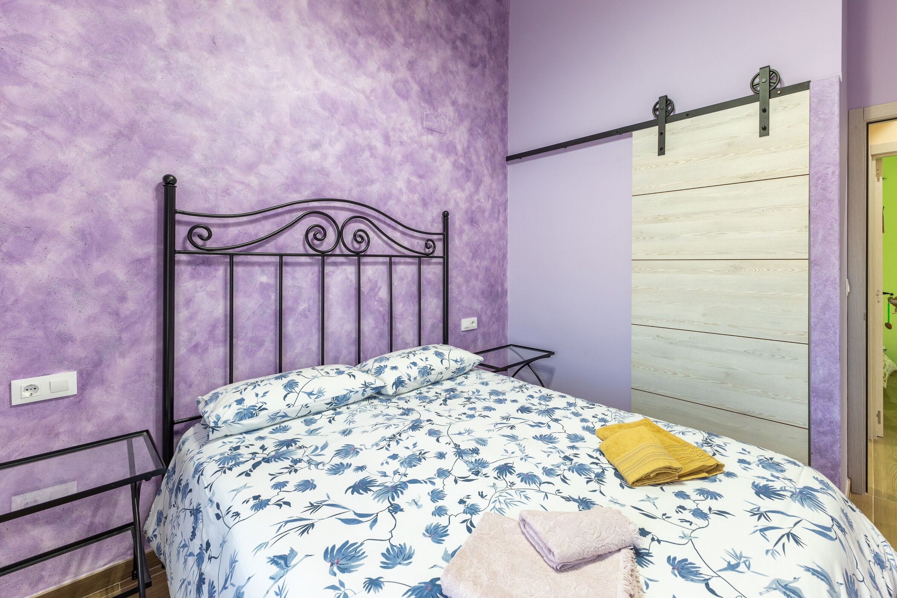 Un dormitorio con una pared morada, un cabecero de cama de metal, una colcha de cama con estampado floral y una puerta corredera de madera.