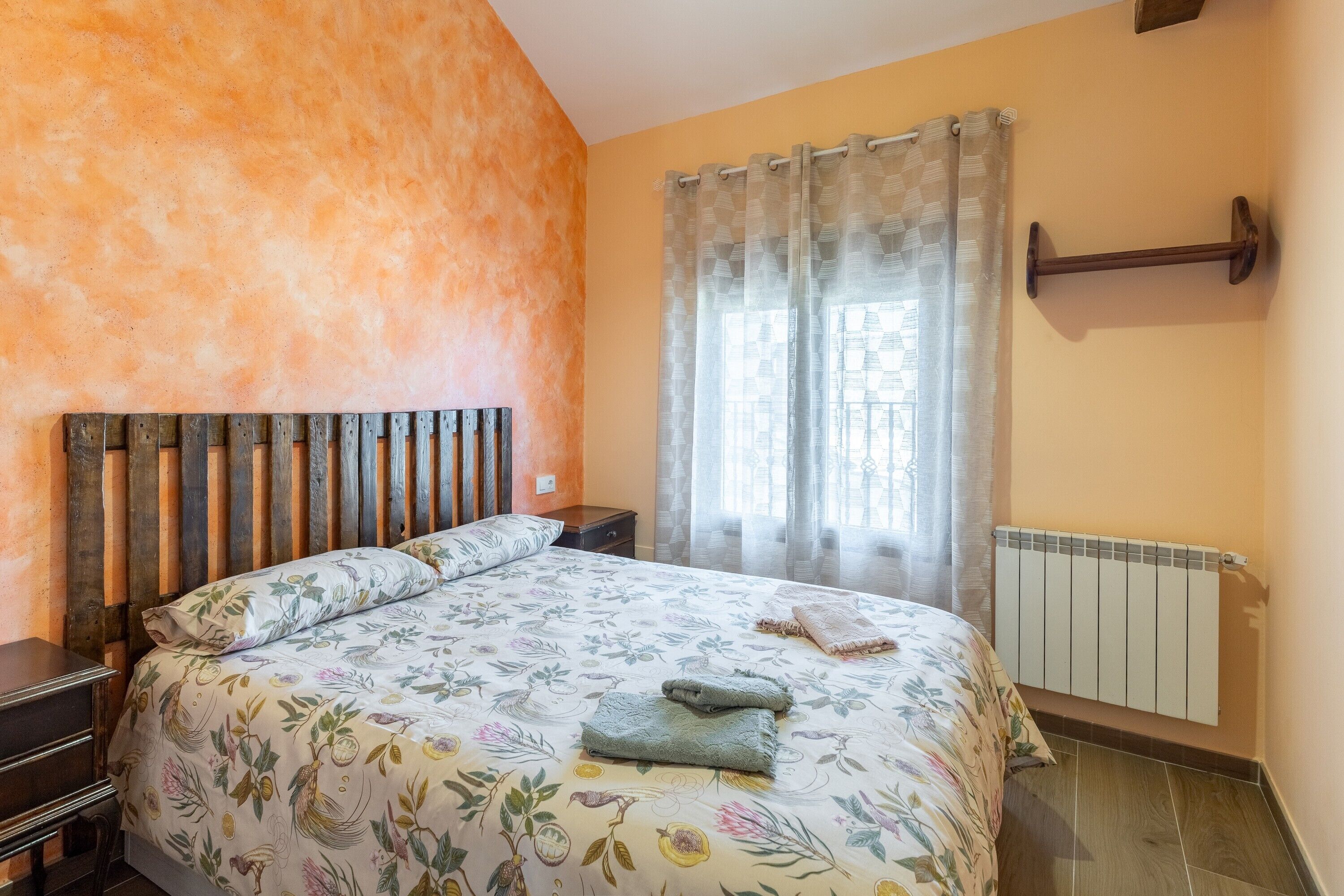 Un dormitorio con cabecera de madera, colcha estampada y radiador, además de una ventana con cortinas.