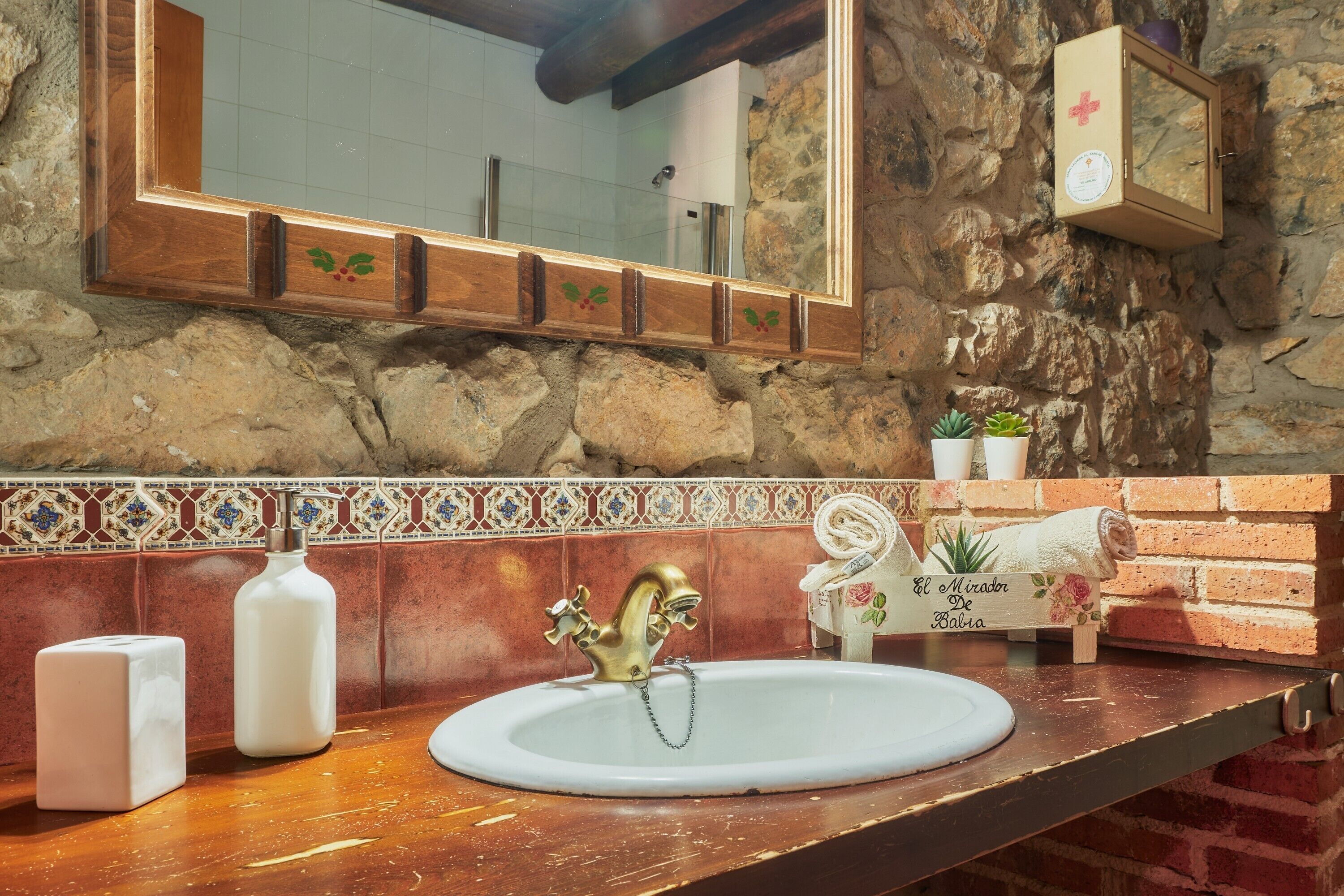 Un baño con pared de piedra, tocador de madera, lavamanos blanco con grifo dorado y un espejo sobre él.
