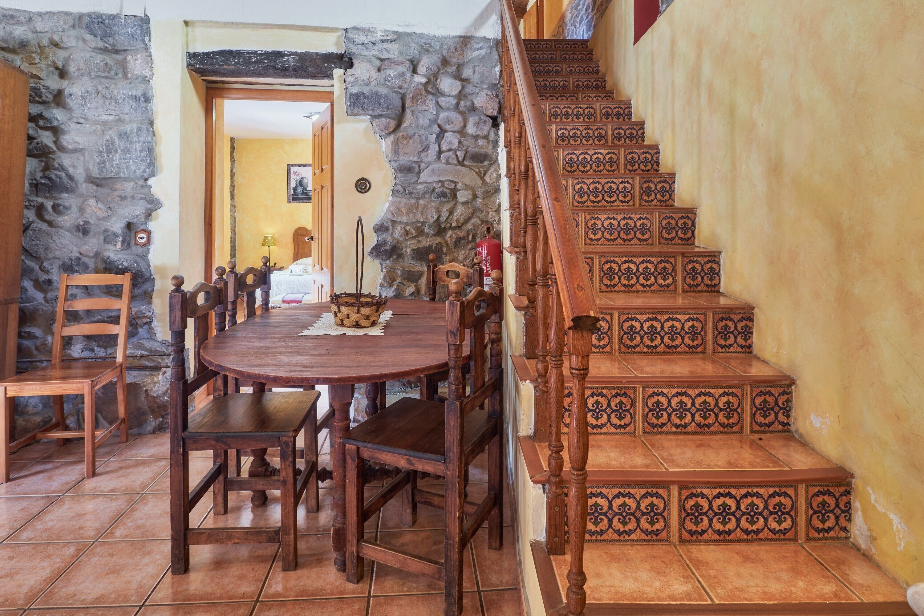 Un comedor con mesa y sillas de madera, un muro de piedra y una escalera con baldosas estampadas.