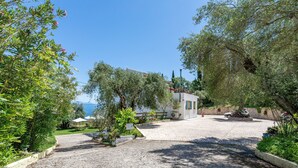 A paved courtyard with a small building, surrounded by trees and a view of the sea. - Room 'Two Bedroom' with Private Terrace, Wi-Fi and Air Conditioning (Corfu)