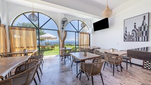 A spacious dining area with large windows, wicker furniture, and a flat-screen TV mounted on the wall. - Room 'Two Bedroom' with Private Terrace, Wi-Fi and Air Conditioning (Corfu)