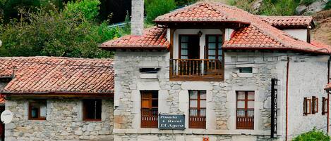 A stone building with a red-tiled roof, multiple windows, and a sign indicating a rural location.