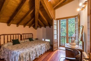 A bedroom with a wooden ceiling, a bed with a floral bedspread, a desk with a vase, and a view of greenery through a window.