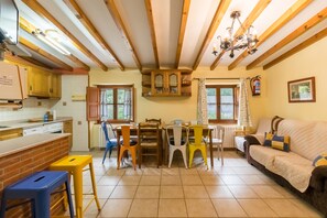 A kitchen with wooden beams, a dining area with chairs, a sofa, and a window with curtains.