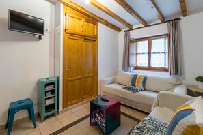 A living room with a sofa, a television mounted on the wall, a wooden wardrobe, a window with curtains, and a small side table.
