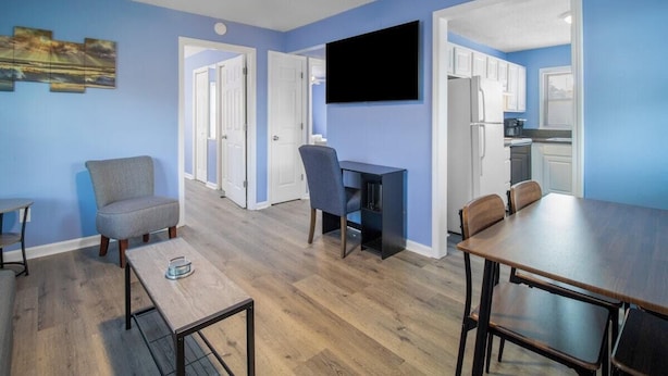 A modern living room with a blue wall, wooden flooring, a ceiling fan, a flat-screen TV, a dining table with chairs, and a small seating area with a chair and a side table.