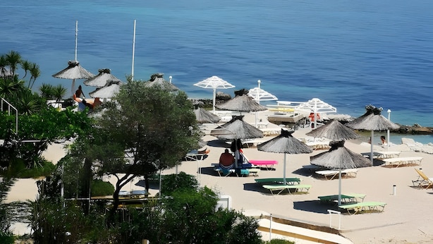 A beach with thatched umbrellas and lounge chairs, a clear view of the sea, and a hilly landscape in the background.