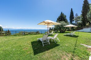 A grassy outdoor area with white lounge chairs and umbrellas, a wooden fence, and a view of the sea and mountains.