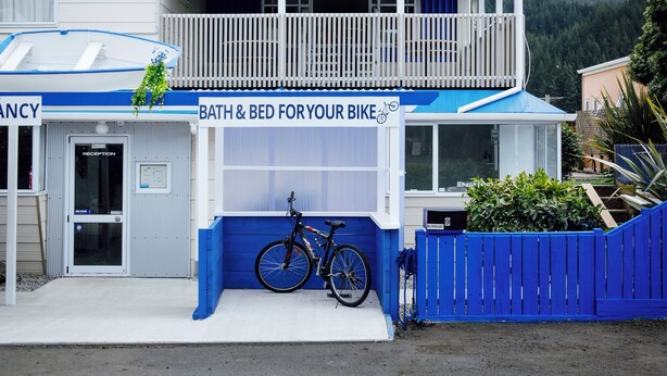 A two-story building with a sign reading "The Boathouse Lodge" and amenities listed such as family rooms, shared dorms, private rooms, in-room bike storage, and free breakfast. - The Boathouse Lodge (Picton)