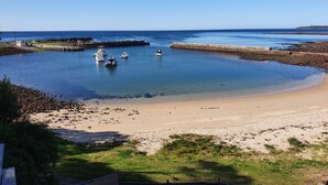 Escena costera con una playa arenosa, agua tranquila y barcos anclados cerca de un rompeolas.