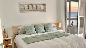 Un dormitorio con una cama grande, un ventilador de techo, vista al océano a través de un ventanal y un letrero en la pared que dice 'BEACH'.