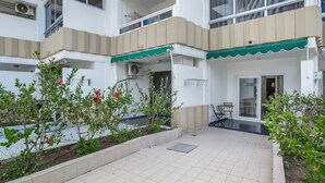 Un edificio residencial de varios pisos con un balcón, un pequeño patio con mesa y sillas, y un jardín con plantas en flor.