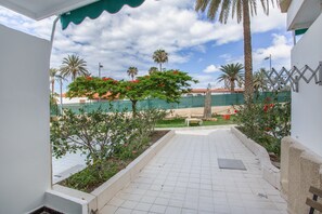 Un patio dallé avec un banc, un arbre aux fleurs rouges et une vue sur un espace clos planté de palmiers.