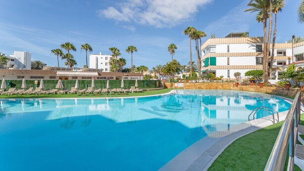 Una piscina con sillones de descanso y sombrillas, rodeada de palmeras y edificios.