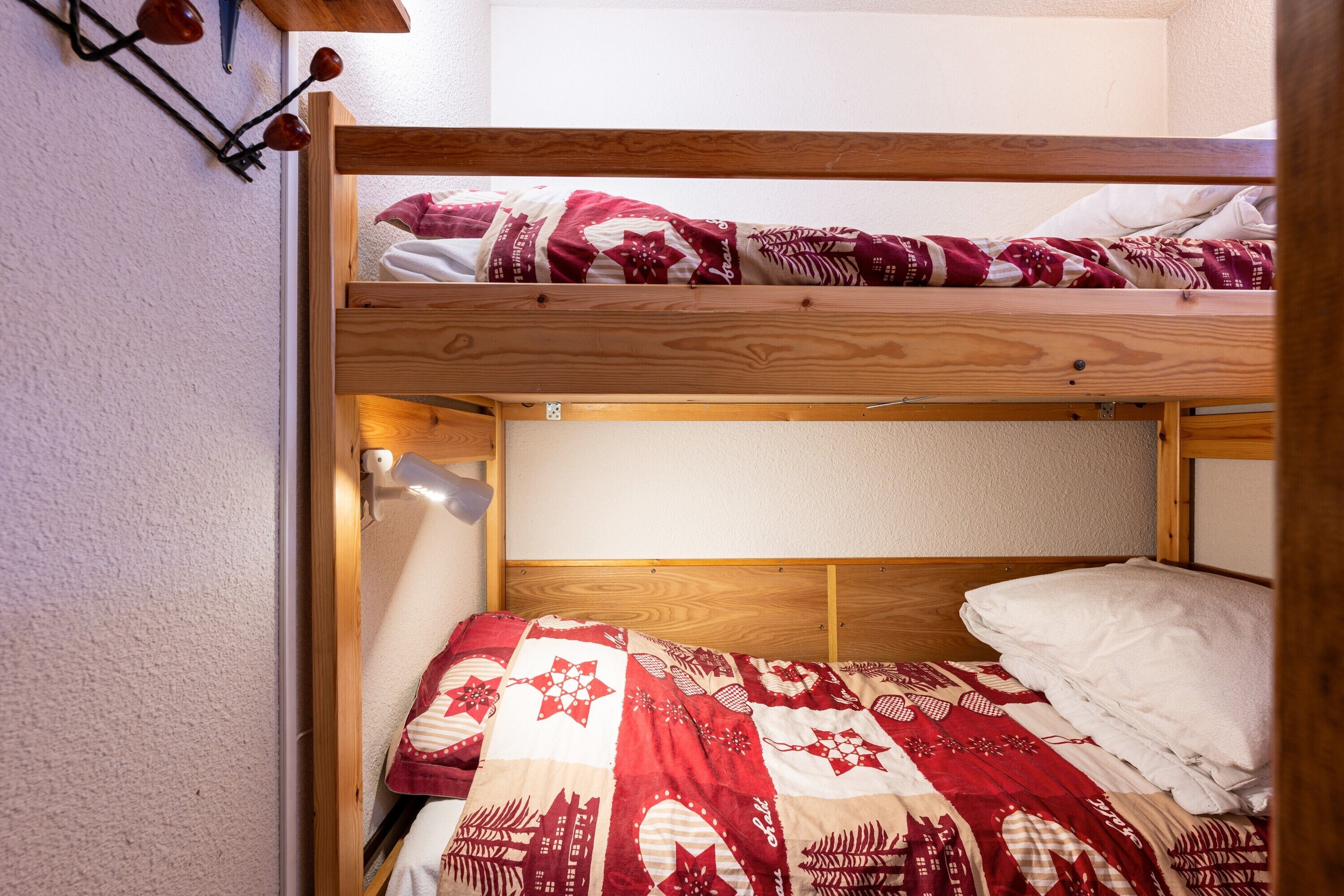 A bunk bed with a red and white quilt, a wooden headboard, and a wall-mounted light fixture.