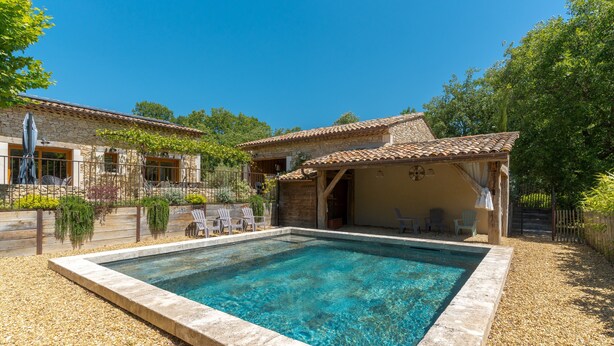 Una piscina con borde de piedra, un porche cubierto y un edificio de piedra con tejado de baldosas.