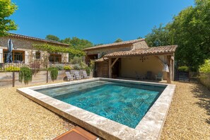 Una piscina con borde de piedra, un porche cubierto y un edificio de piedra con tejado de baldosas.