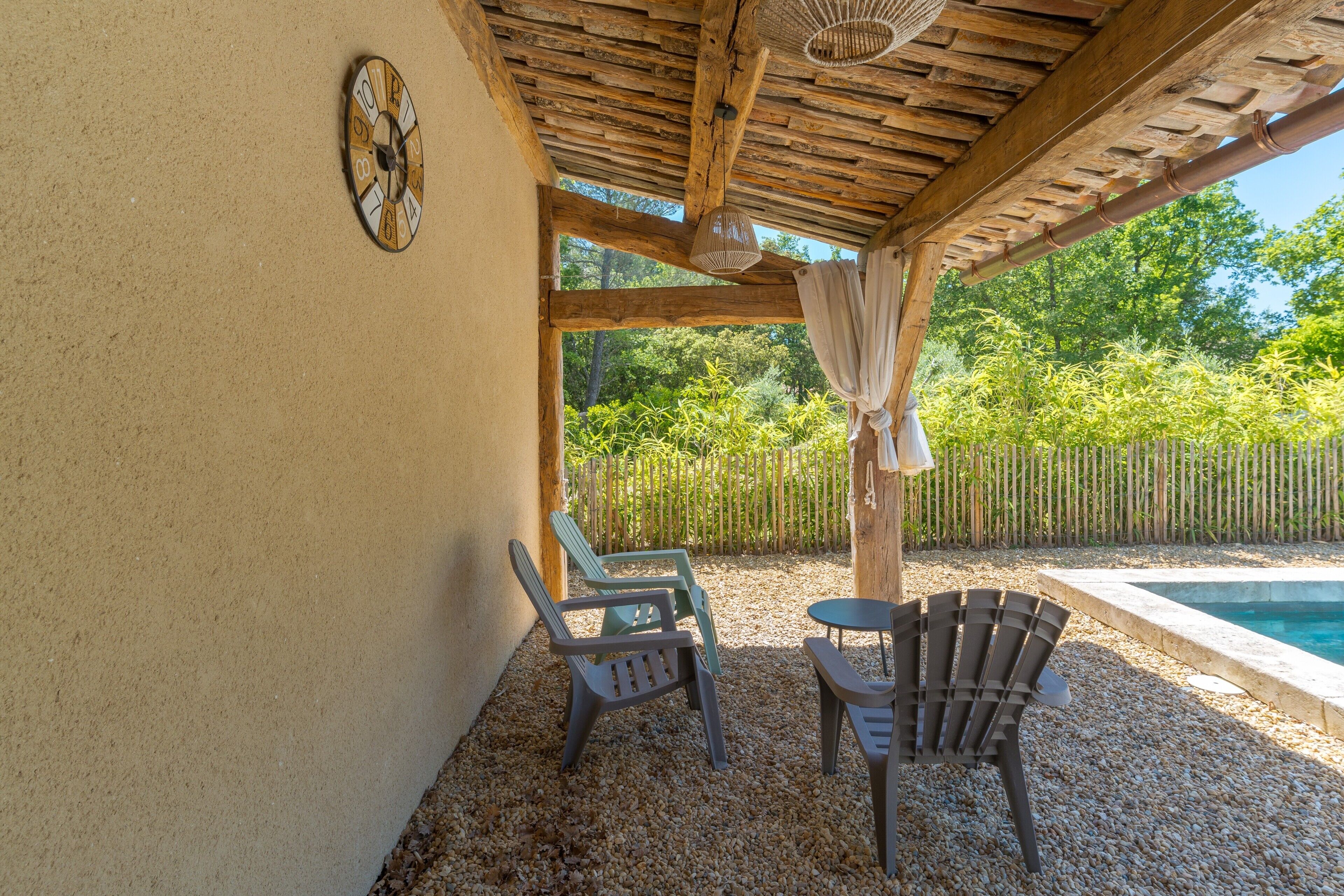 Un área de estar al aire libre con techo, sillas y una mesa, un reloj en la pared, y vistas a una piscina y zonas verdes.