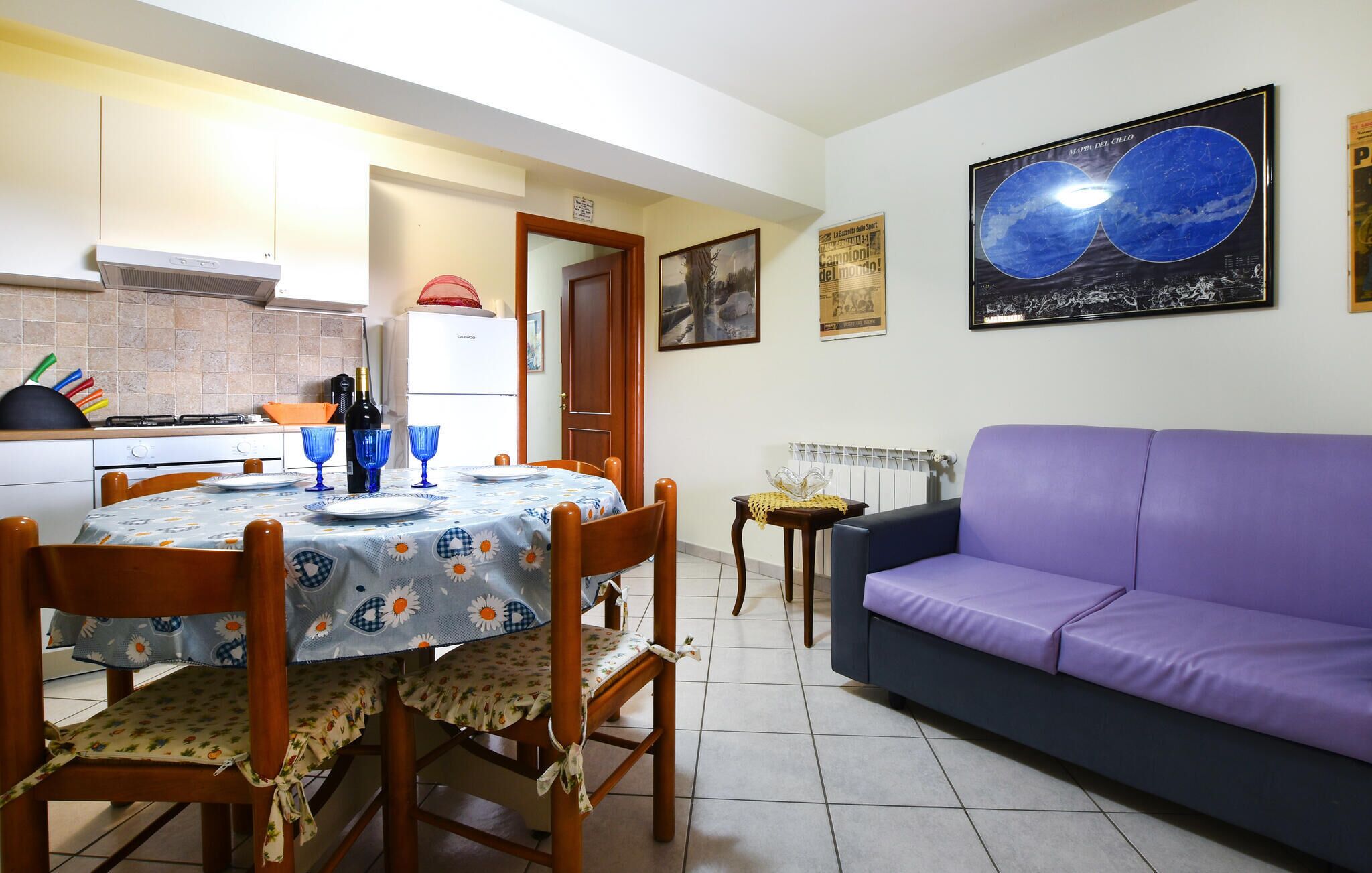 A dining area with a table set for two, a purple sofa, and a kitchen in the background.