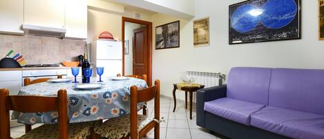 A dining area with a table set for two, a purple sofa, and a kitchen in the background.