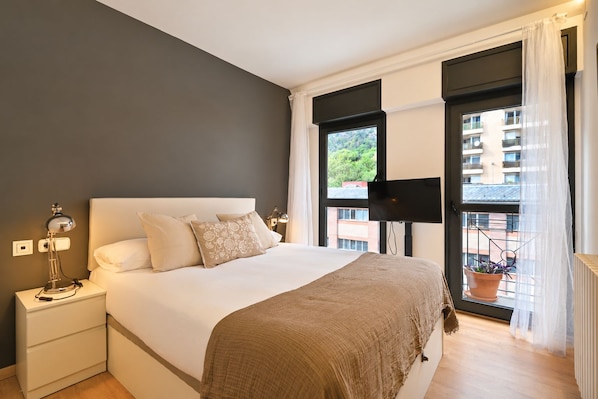A bedroom with a large bed, bedside lamps, a flat-screen TV, and a window with a view of buildings and greenery.