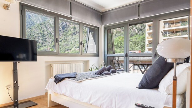 Quarto moderno com uma cama grande, uma televisão e vista para uma varanda com montanhas ao fundo.