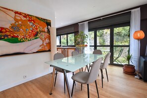 A dining area with a glass table and chairs, a large painting on the wall, and a view of greenery through large windows.