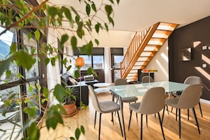 A modern living room with a glass dining table, comfortable seating, a staircase with wooden railings, and a view through large windows.