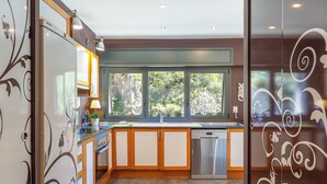 Uma cozinha moderna com armários de madeira, uma máquina de lavar louça em aço inoxidável e uma grande janela com vista para árvores.