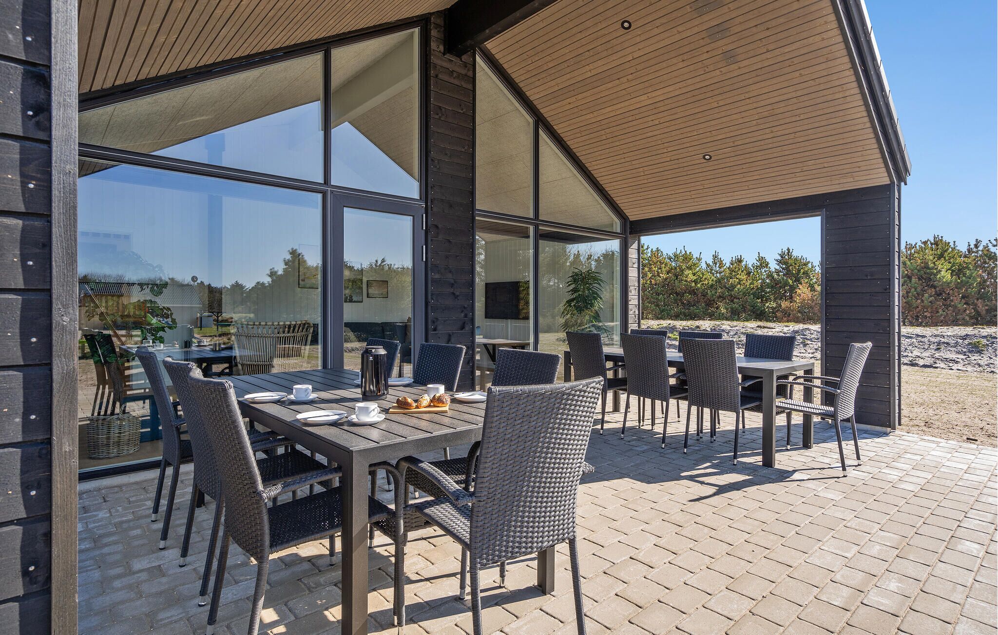 An outdoor dining area with a modern design, featuring a large table and multiple chairs, set on a paved patio with a view of the surrounding landscape.