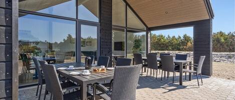 An outdoor dining area with a modern design, featuring a large table and multiple chairs, set on a paved patio with a view of the surrounding landscape.