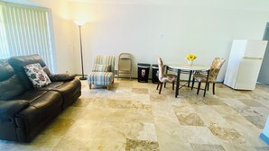 A living room with a dark leather sofa, a small round table with chairs, a standing lamp, and a white refrigerator. - Guest Suite East Boca! Near Beach & Restaurants! Near FAU! Close to I-95! (Boca Raton)