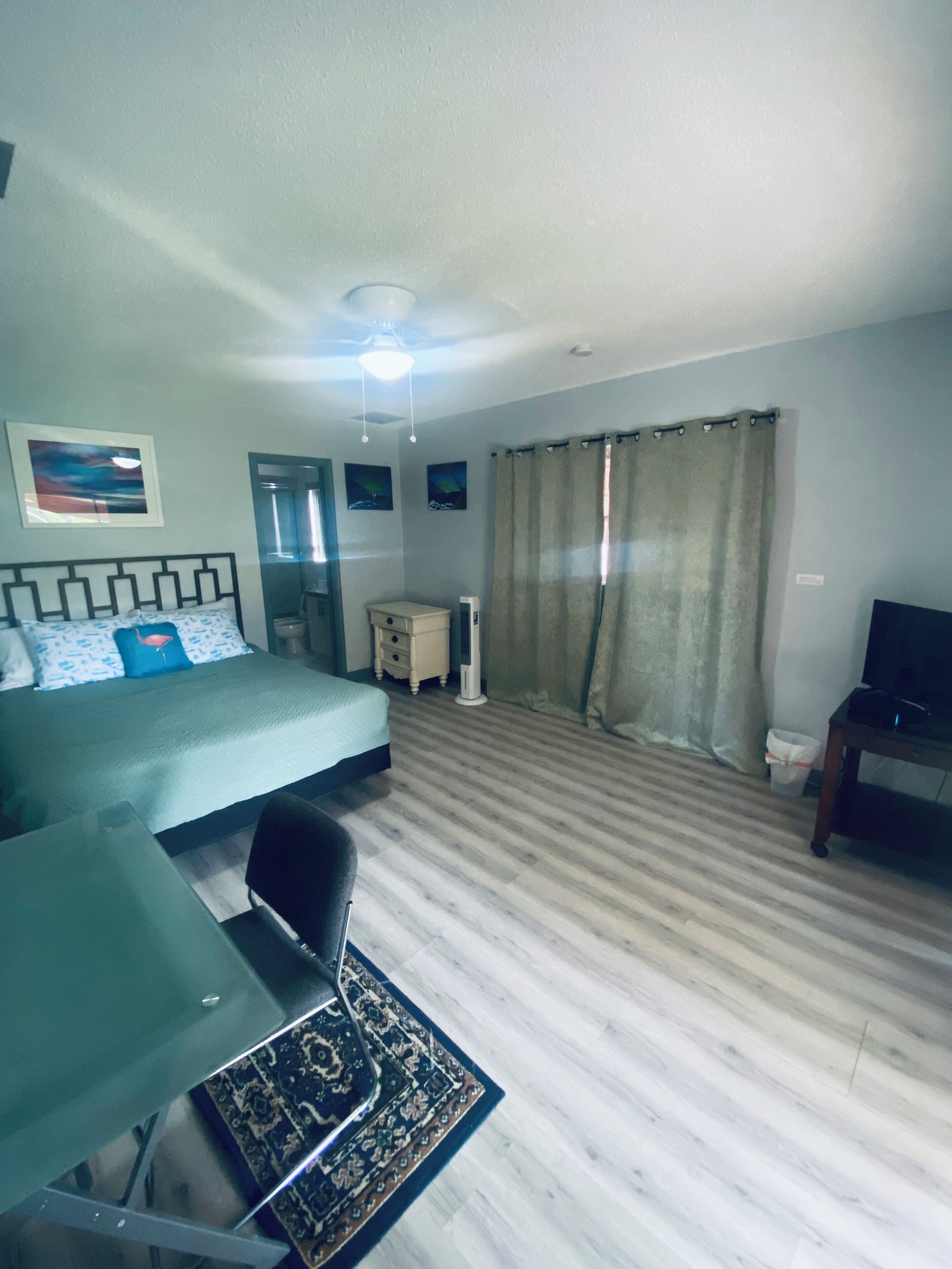 Ein Hotelzimmer mit Bett, Schreibtisch, Stuhl, Fernseher, Deckenventilator und einem Fenster mit Vorhängen.