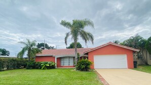 A single-story house with a red facade, a white garage door, and a well-maintained lawn. - Guest Suite East Boca! Near Beach & Restaurants! Near FAU! Close to I-95! (Boca Raton)