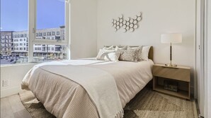 Een slaapkamer met een groot raam, een bed met een beige deken, een nachtkastje met een lamp, en een kunstwerk aan de muur.