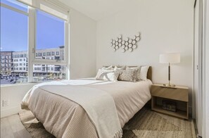 A bedroom with a large window, a bed with a beige blanket, a nightstand with a lamp, and a wall art piece. - New Studio at Stanley Marketplace (Aurora)