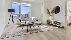 Een moderne woonkamer met een bank, salontafel en vloerlamp. Er is een groot raam met uitzicht op de stad, een witte console tafel en een decoratieve vaas met pluimveedergras.