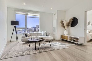 A modern living room with a sofa, coffee table, and floor lamp. There is a large window with a city view, a white console table, and a decorative vase with pampas grass. - New Studio at Stanley Marketplace (Aurora)