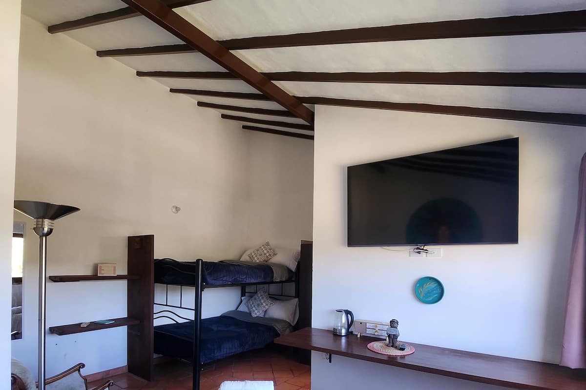 A bedroom with a bed, a desk, a TV mounted on the wall, and a bunk bed in the background.