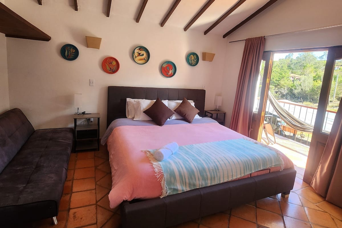 A bedroom with a bed, a sofa, a nightstand, and a window with a view of greenery.