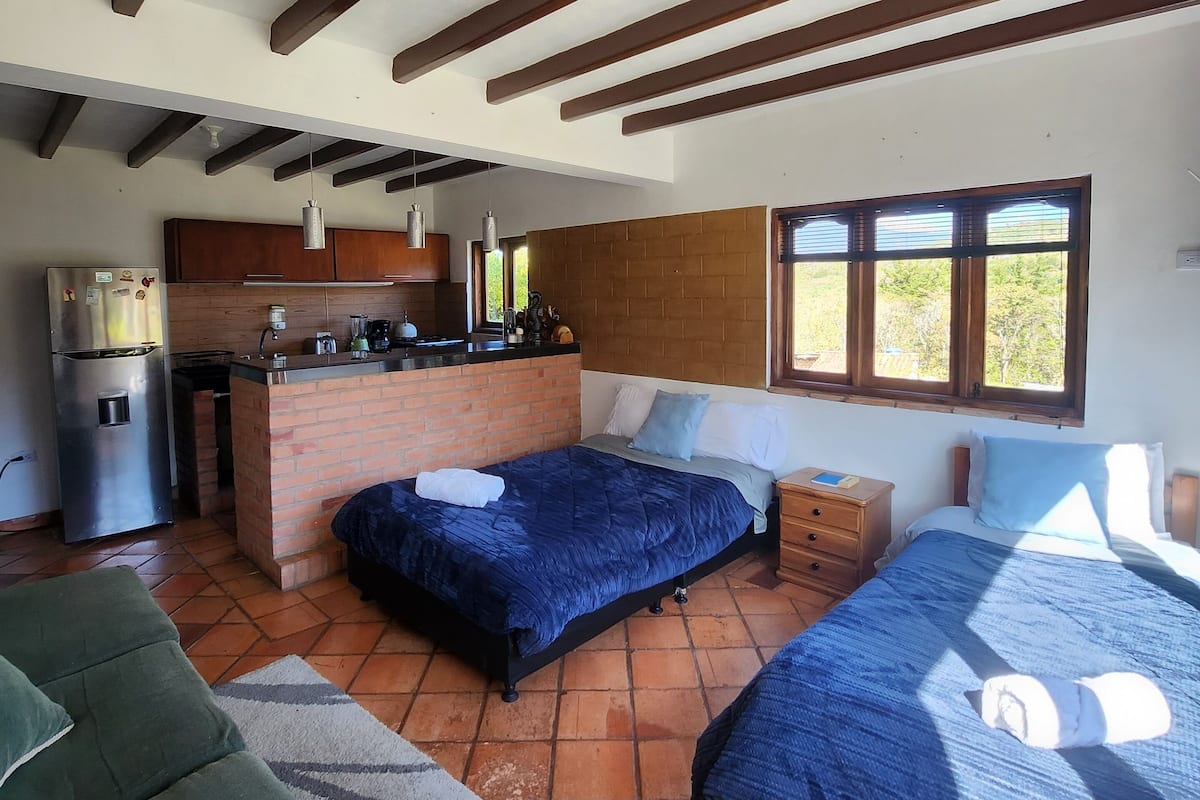 A compact room with a kitchenette, two beds, a sofa, and a window with blinds.