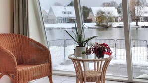 A room with a wicker chair and a small table with a plant and flowers, overlooking a snowy river with houses in the background.