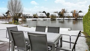 A lakeside patio with metal chairs and tables, overlooking a calm lake and residential buildings with snow-covered roofs.